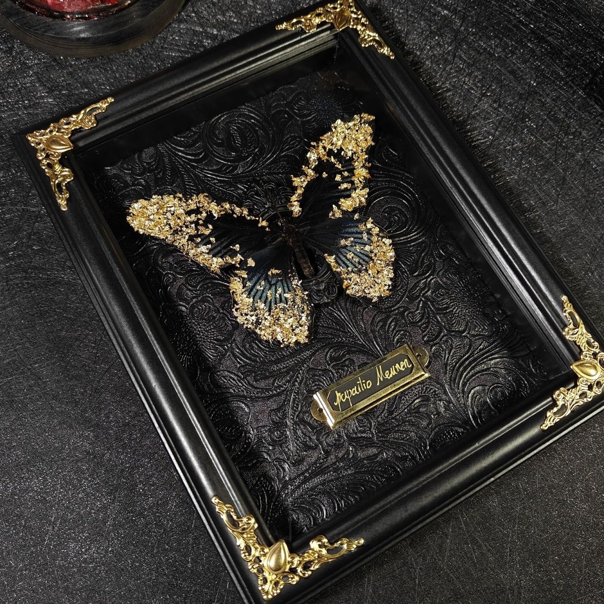 InsectArts framed Papilio Memnon butterfly specimen. Black wings embellished with gold leaf flakes mounted on a dark embossed baroque background. Features a vintage brass nameplate and black frame with ornate gold corners. Premium taxidermy art for dark academia decor.